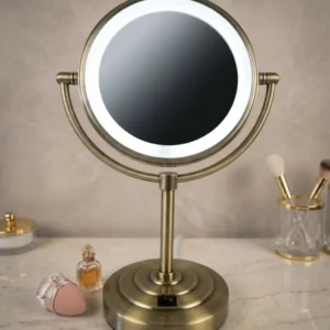 An illuminated vanity mirror with a round LED ring and dual-angle stand, shown on a marble countertop with cosmetic items in a bathroom setting.